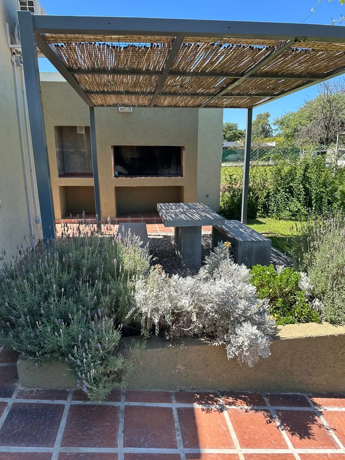 Parrillero de material con pérgola de caña, mesa y bancos de hormigón junto al jardín